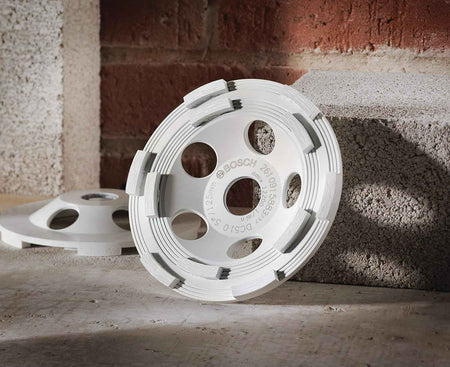 A product shot of the white Bosch diamond cup wheel propped against a cinder block in an industrial setting with another wheel visible in the background.