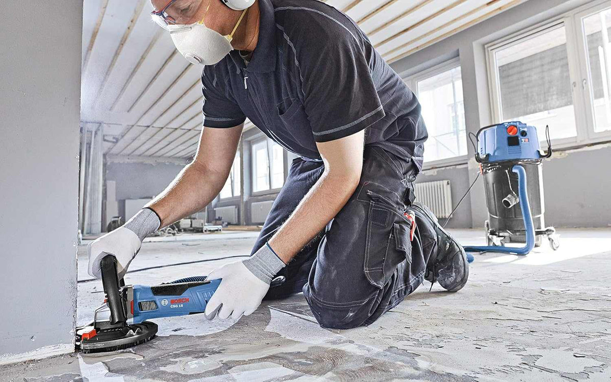An action shot of an operator wearing safety gear and kneeling on a concrete floor while using a Bosch grinder with a dust shroud and vacuum attachment to grind along the edge of a wall.