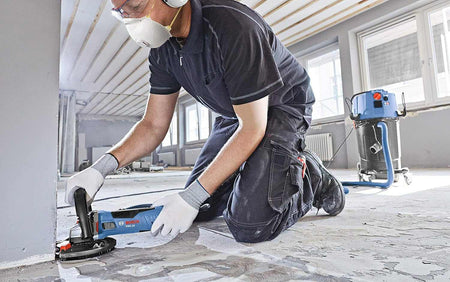 An action shot of an operator wearing safety gear and kneeling on a concrete floor while using a Bosch grinder with a dust shroud and vacuum attachment to grind along the edge of a wall.