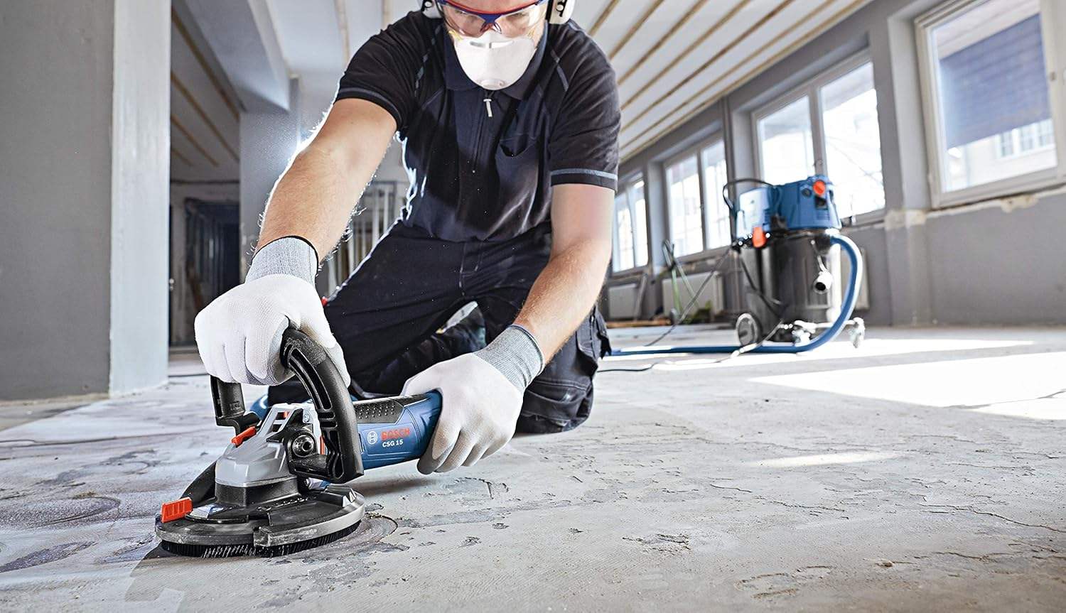 A front-view action shot of an operator wearing protective equipment while using a Bosch surfacing grinder to level a concrete floor in an open industrial room.