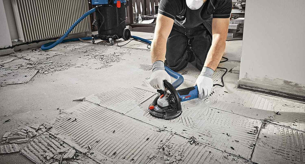 An action shot of an operator using a Bosch grinder with a vacuum-connected dust shroud to remove old adhesive from a concrete floor, with a blue industrial vacuum visible in the background.