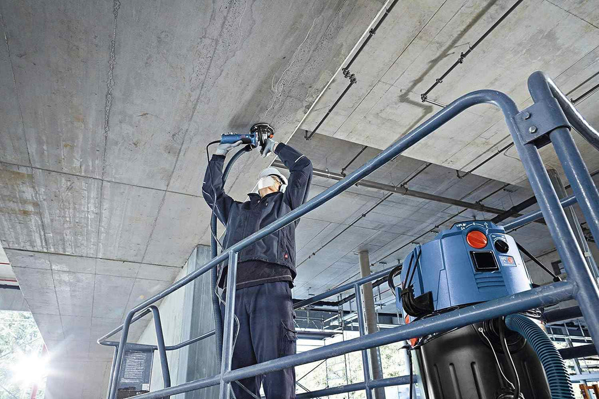 An action shot of an operator wearing a hard hat and safety gear while standing on a scaffold to grind the underside of a concrete ceiling using a Bosch grinder connected to a dust extraction system.