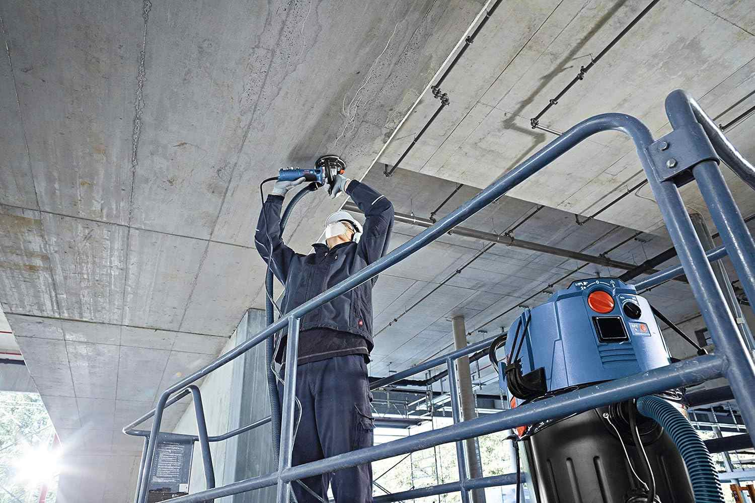 An action shot of an operator wearing a hard hat and safety gear while standing on a scaffold to grind the underside of a concrete ceiling using a Bosch grinder connected to a dust extraction system.