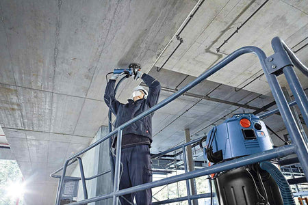 An action shot of an operator wearing a hard hat and safety gear while standing on a scaffold to grind the underside of a concrete ceiling using a Bosch grinder connected to a dust extraction system.