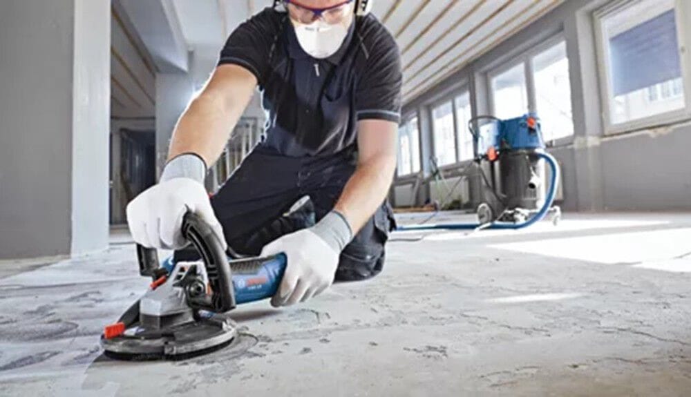 A construction worker on his knees wearing a dust mask, safety glasses, and protective gloves handles a handheld Bosch angle grinder fitted with a dust extraction shroud and a diamond cup wheel to flatten a rough indoor concrete floor while a blue dust extractor vacuum runs in the background.