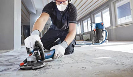 A construction worker on his knees wearing a dust mask, safety glasses, and protective gloves handles a handheld Bosch angle grinder fitted with a dust extraction shroud and a diamond cup wheel to flatten a rough indoor concrete floor while a blue dust extractor vacuum runs in the background.