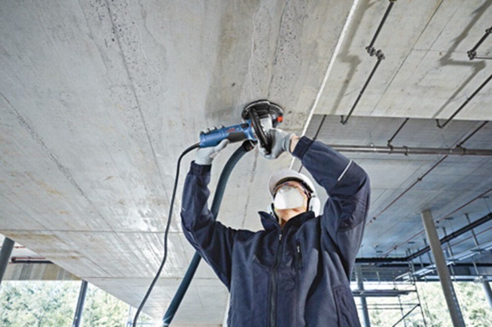An overhead action shot of a tradesperson wearing a white hard hat, face mask, and safety earmuffs using a shrouded Bosch concrete surface grinder to level an unfinished concrete ceiling slab with a vacuum hose attached for dust collection.