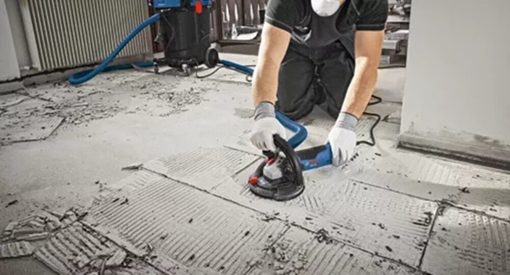 A flooring installer working on a renovation project uses an unthreaded 7/8-inch arbor diamond grinding cup wheel on a Bosch angle grinder to clear away hard thinset mortar ridges and prepare the structural concrete subfloor for new tile installation.