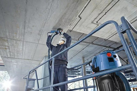 A commercial contractor standing safely on a scaffolding structure utilizes a 4-1/2 inch diamond cup wheel system connected to a heavy-duty Bosch vacuum cleaner unit to plane down high spots on a rough overhead concrete slab structure.