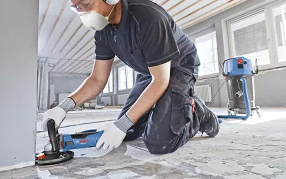 A close-up view of a construction professional working flush against an interior wall corner, utilizing a high-speed Bosch diamond grinding disc wheel with a protective brush-ring dust shroud to level down rough aggregate concrete surfaces safely.
