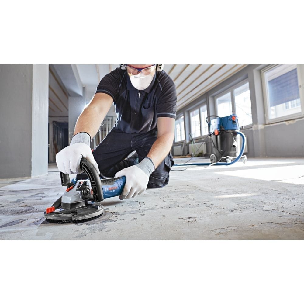 An indoor jobsite application photo showing a professional contractor on his knees utilizing a shrouded Bosch concrete surface grinder connected to a blue dust extraction vacuum to safely strip old paint coatings and level an uneven commercial concrete floor.