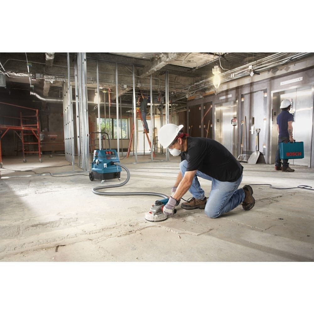 A wide-angle construction site view showing a worker wearing a white hard hat and safety dust mask as he operates a hand grinder with a dust shroud on a wide concrete slab foundation, utilizing the Bosch 5-inch diamond cup wheel system connected to a matching blue dust collection extractor vacuum unit to remove surface coatings.