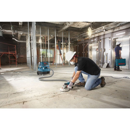 A wide-angle construction site view showing a worker wearing a white hard hat and safety dust mask as he operates a hand grinder with a dust shroud on a wide concrete slab foundation, utilizing the Bosch 5-inch diamond cup wheel system connected to a matching blue dust collection extractor vacuum unit to remove surface coatings.