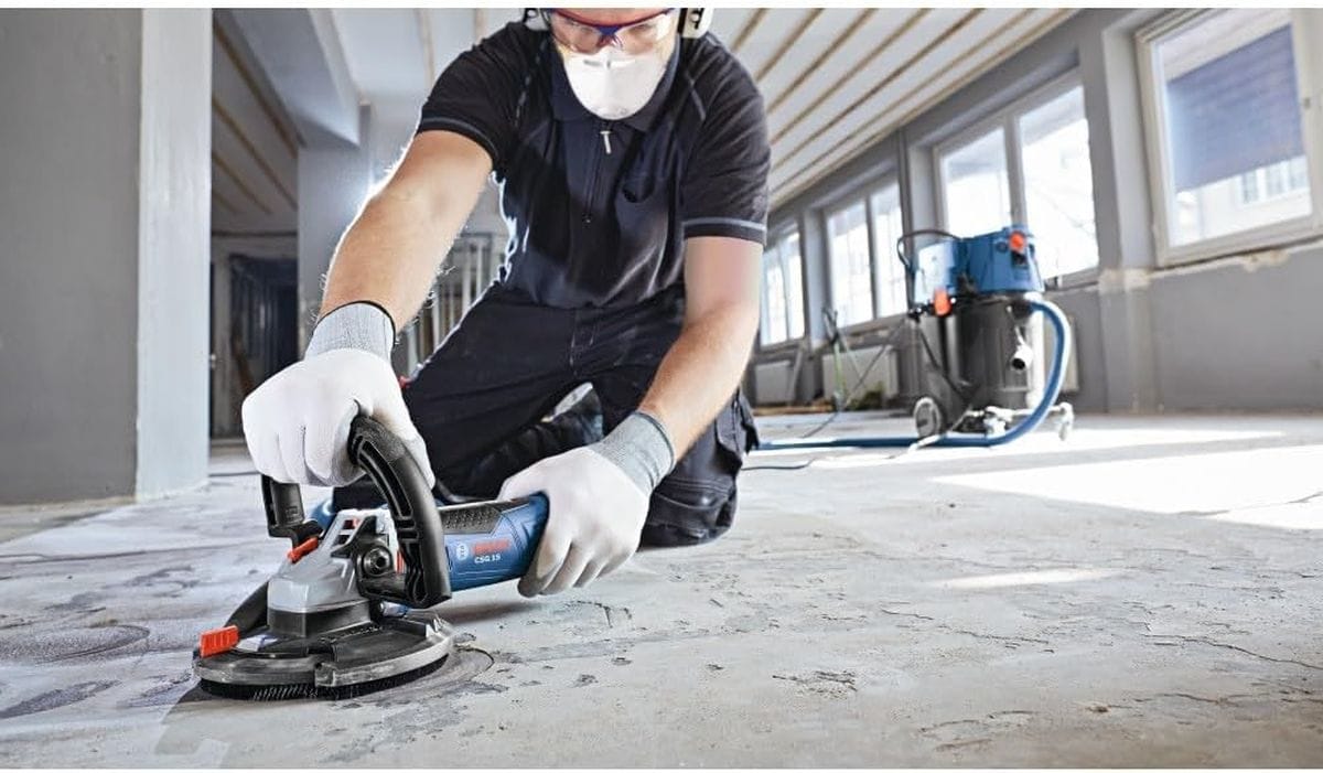 An indoor jobsite application shot of a flooring installer on his knees wearing safety equipment, utilizing a handheld Bosch concrete surface planer equipped with a vacuum-connected extraction dust shroud to level down a rough, uneven concrete subfloor layout.