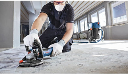 An indoor jobsite application shot of a flooring installer on his knees wearing safety equipment, utilizing a handheld Bosch concrete surface planer equipped with a vacuum-connected extraction dust shroud to level down a rough, uneven concrete subfloor layout.