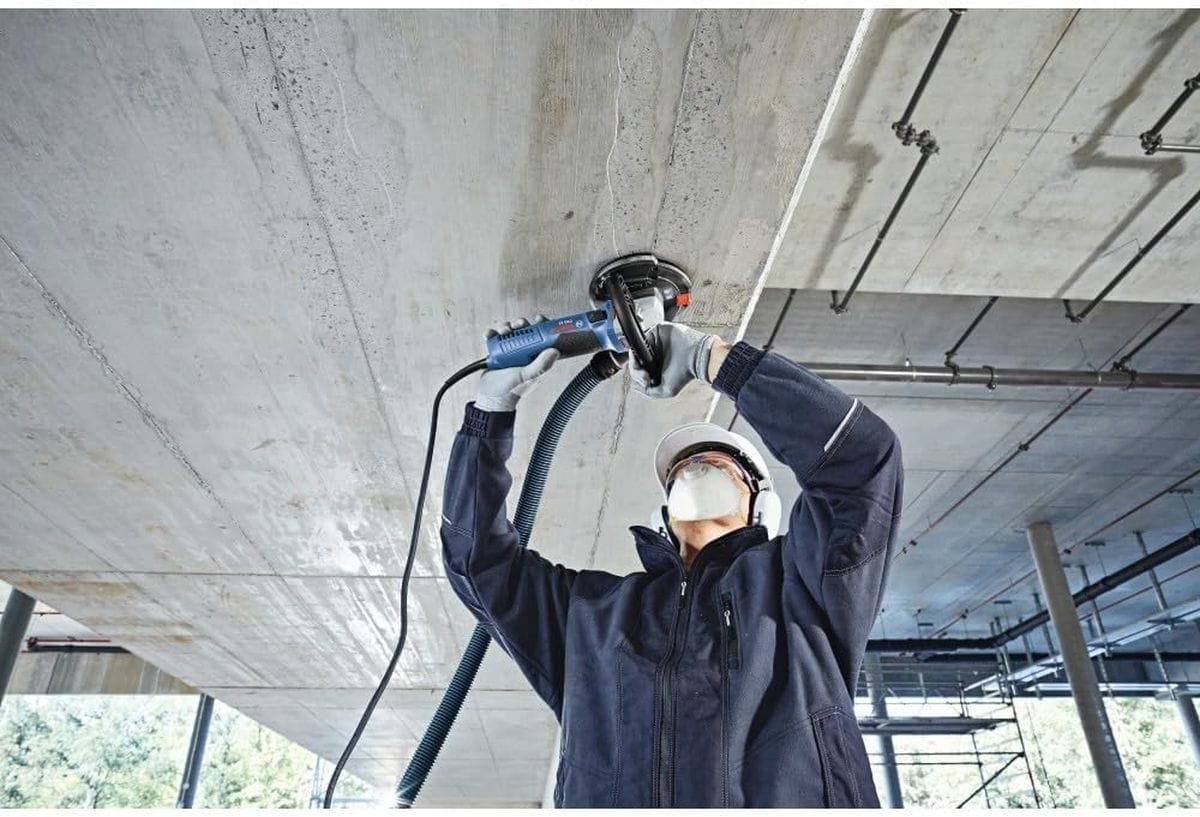 An overhead action shot of a commercial contractor wearing a white hard hat, face protection mask, and ear defenders while using a shrouded Bosch hand grinder to flatten and smooth high spots along a structural poured concrete ceiling slab.