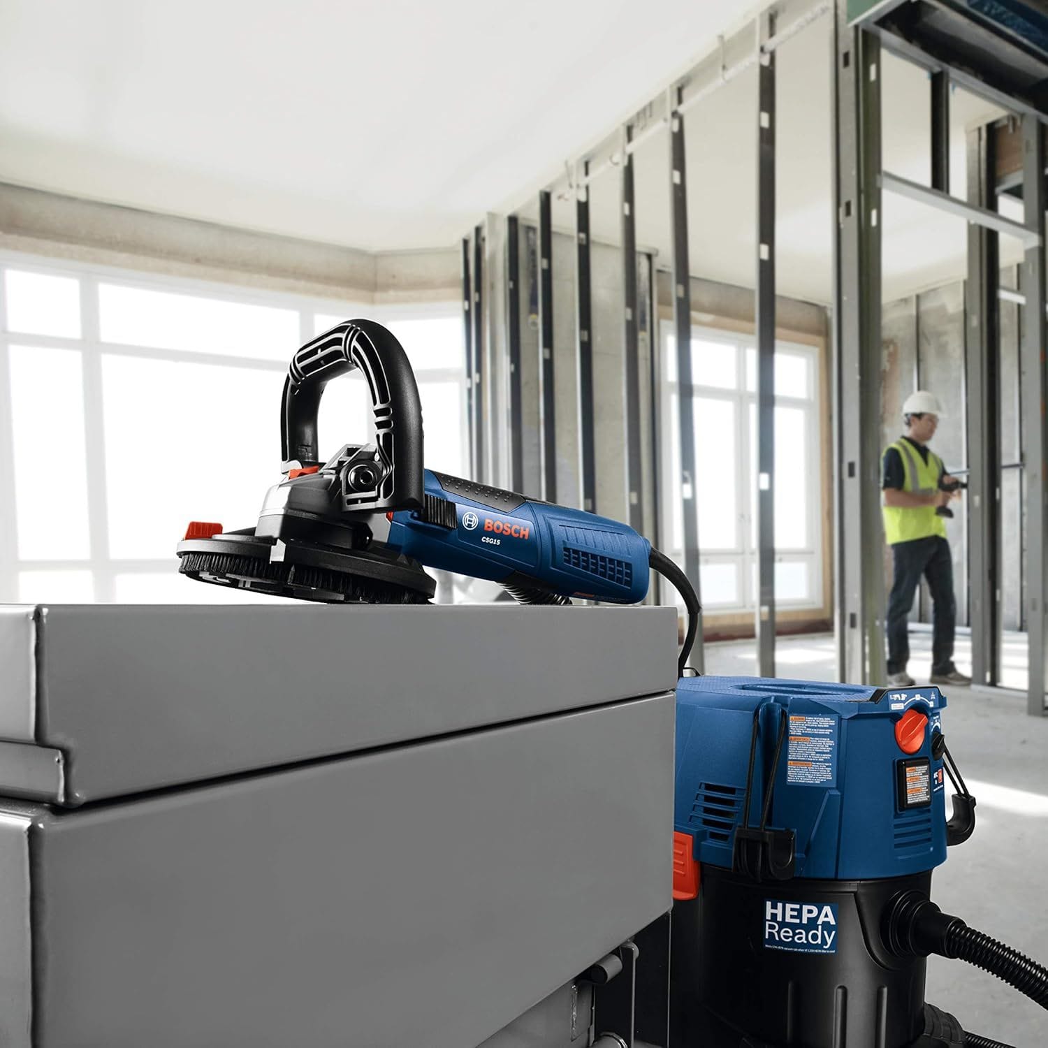 A professional product application layout showing a blue corded Bosch concrete surface tool resting on a metal storage bin box, fully assembled with a protective brush dust shroud and connected via a flexible hose line to an industrial blue Bosch HEPA Ready dust extractor vacuum unit on a commercial construction site.