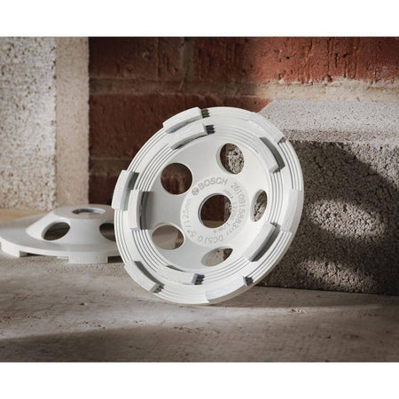 A jobsite layout photo showing a white Bosch diamond grinding cup wheel propped up against a rough concrete masonry block in front of a red brick wall background, highlighting the raised diamond cutting segments and internal unthreaded cooling ports.