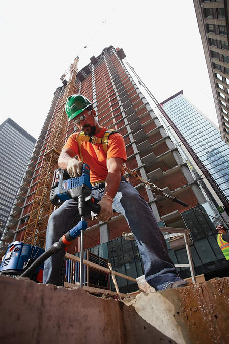 A low-angle view looking up at a commercial ironworker or concrete mason standing against a high-rise building framework, using a cordless Bosch SDS-plus hammer drill with a Speed Clean extraction bit to prepare structural anchor settings on an open-air concrete ledge.