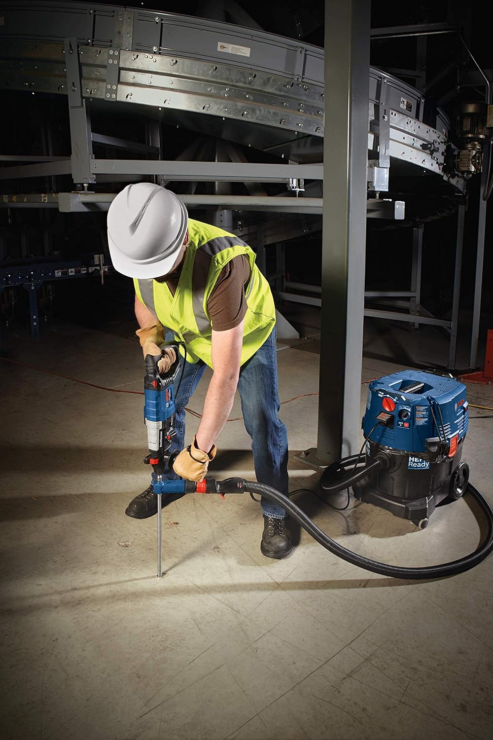 A commercial construction professional wearing a safety vest and hard hat utilizes a heavy-duty rotary hammer equipped with the Bosch DXS2074 extraction bit to drill vertically into a warehouse concrete floor, with the suction hose routing debris straight into a matching blue Bosch dust extractor vacuum.