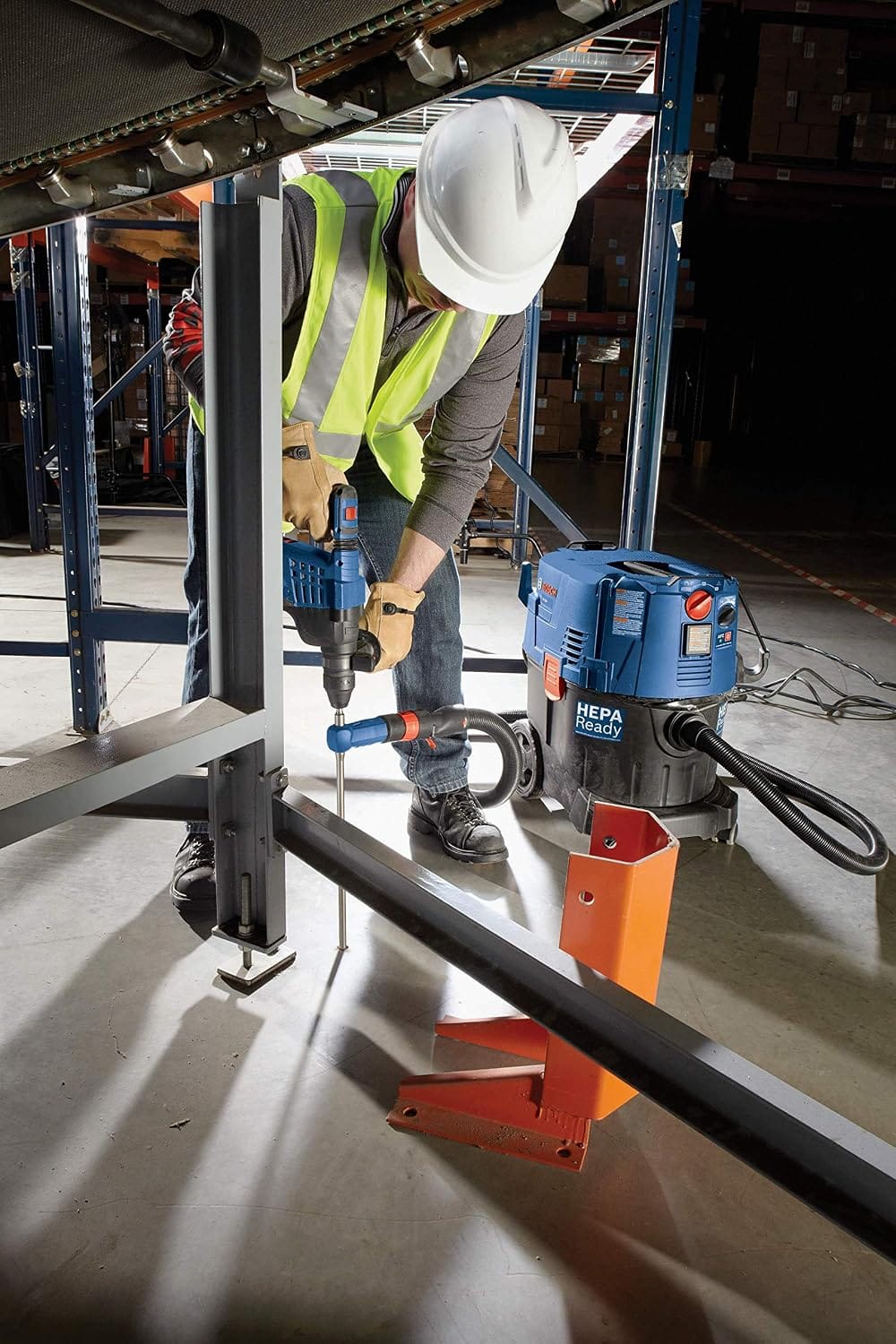 A close-up shot of a trade worker wearing safety gear operating an SDS-plus hammer drill and a Bosch hollow dust extraction bit next to industrial warehouse shelving units, pulling harmful masonry concrete dust away from the hole dynamically through a connected vacuum system.
