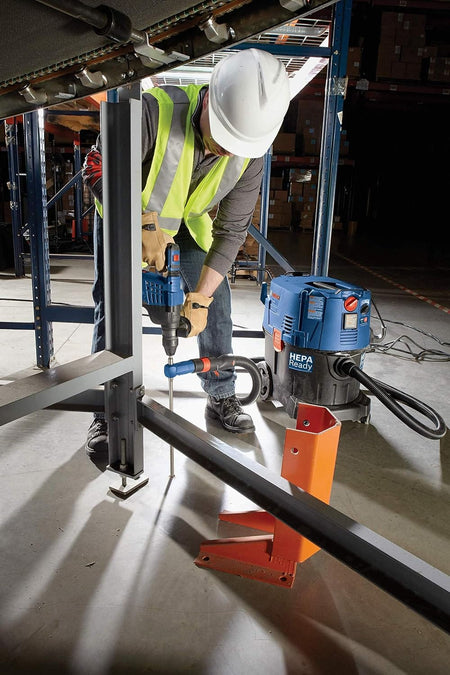 A close-up shot of a trade worker wearing safety gear operating an SDS-plus hammer drill and a Bosch hollow dust extraction bit next to industrial warehouse shelving units, pulling harmful masonry concrete dust away from the hole dynamically through a connected vacuum system.