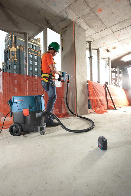 An outdoor construction scene showing a worker wearing a safety harness and green hard hat drilling horizontally into a concrete column with a Bosch dustless hammer drill bit system, while a laser level sits nearby on the jobsite floor to ensure perfect structural alignment.
