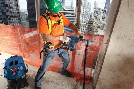 An outdoor construction professional wearing a green hard hat, orange shirt, and full safety harness uses a cordless Bosch rotary hammer drill fitted with the DXS2114 hollow extraction bit to drill into a concrete structural column on an open high-rise building floor, with a blue HEPA-ready vacuum setup extraction system attached.