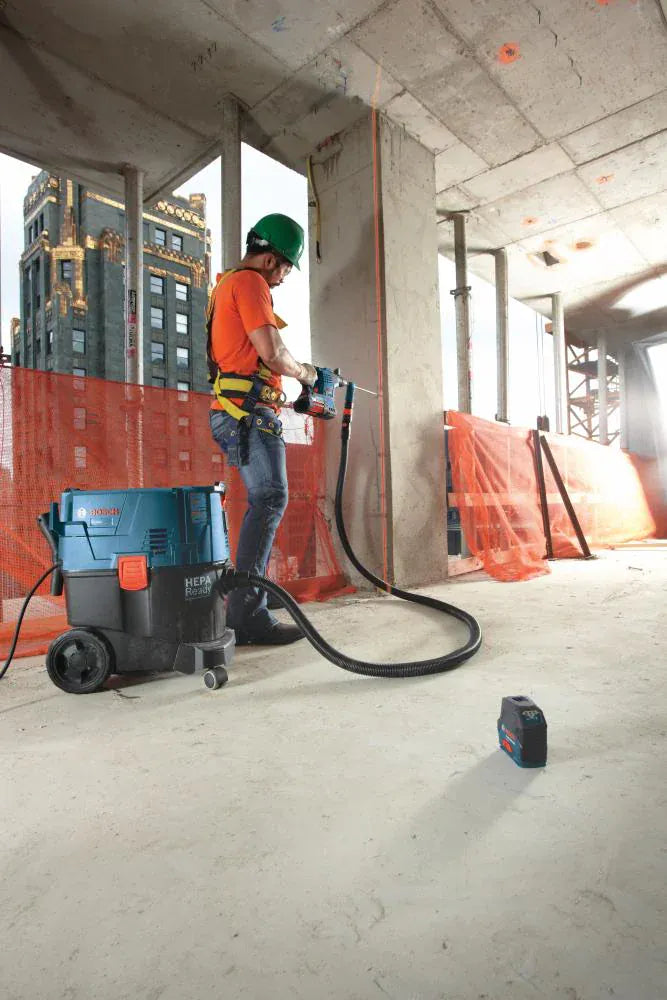 A construction worker operating a cordless SDS-plus rotary hammer with the Bosch hollow dustless extraction bit system to drill horizontally into a concrete support wall, utilizing an adjacent blue Bosch dust extractor vacuum while a compact line laser rests on the concrete floor for alignment.