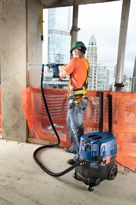An industrial jobsite view showing an ironworker or concrete mason utilizing a Bosch dustless hammer drill bit system to drill a deep anchoring hole into a concrete partition on an upper floor level, showing the vacuum extraction hose routing debris dynamically into a blue HEPA-ready dust extractor vacuum against a city skyline.