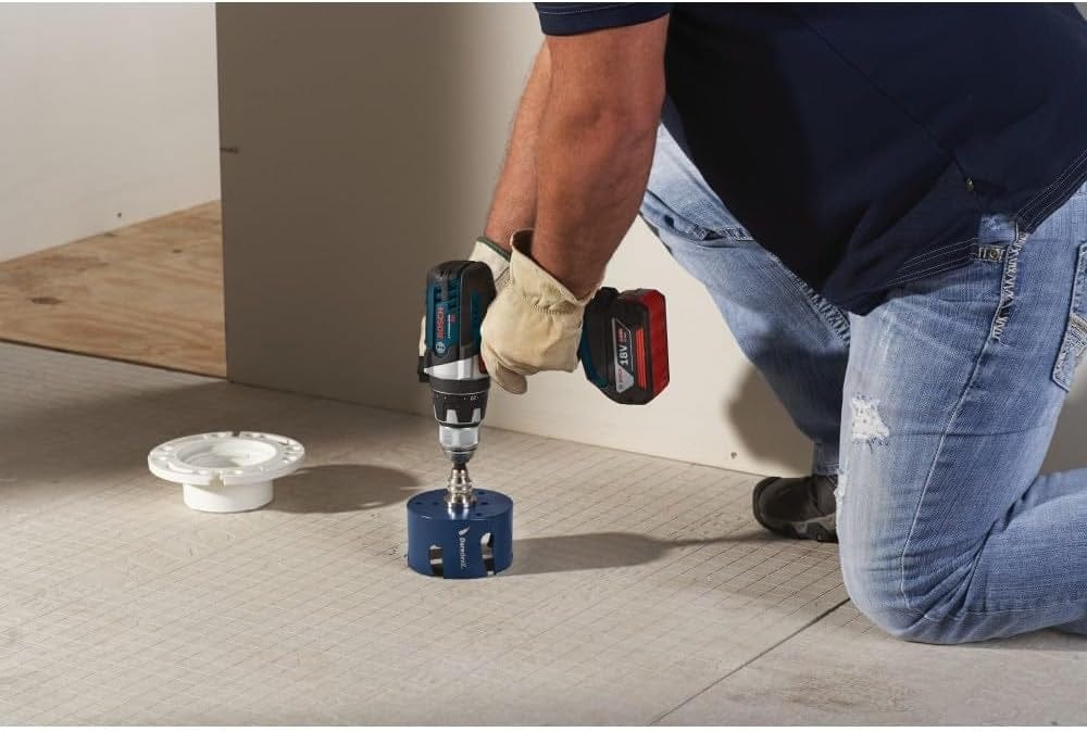 Close-up of a worker using a cordless drill and the blue hole saw to cut a hole in cement board flooring near a white pipe flange.