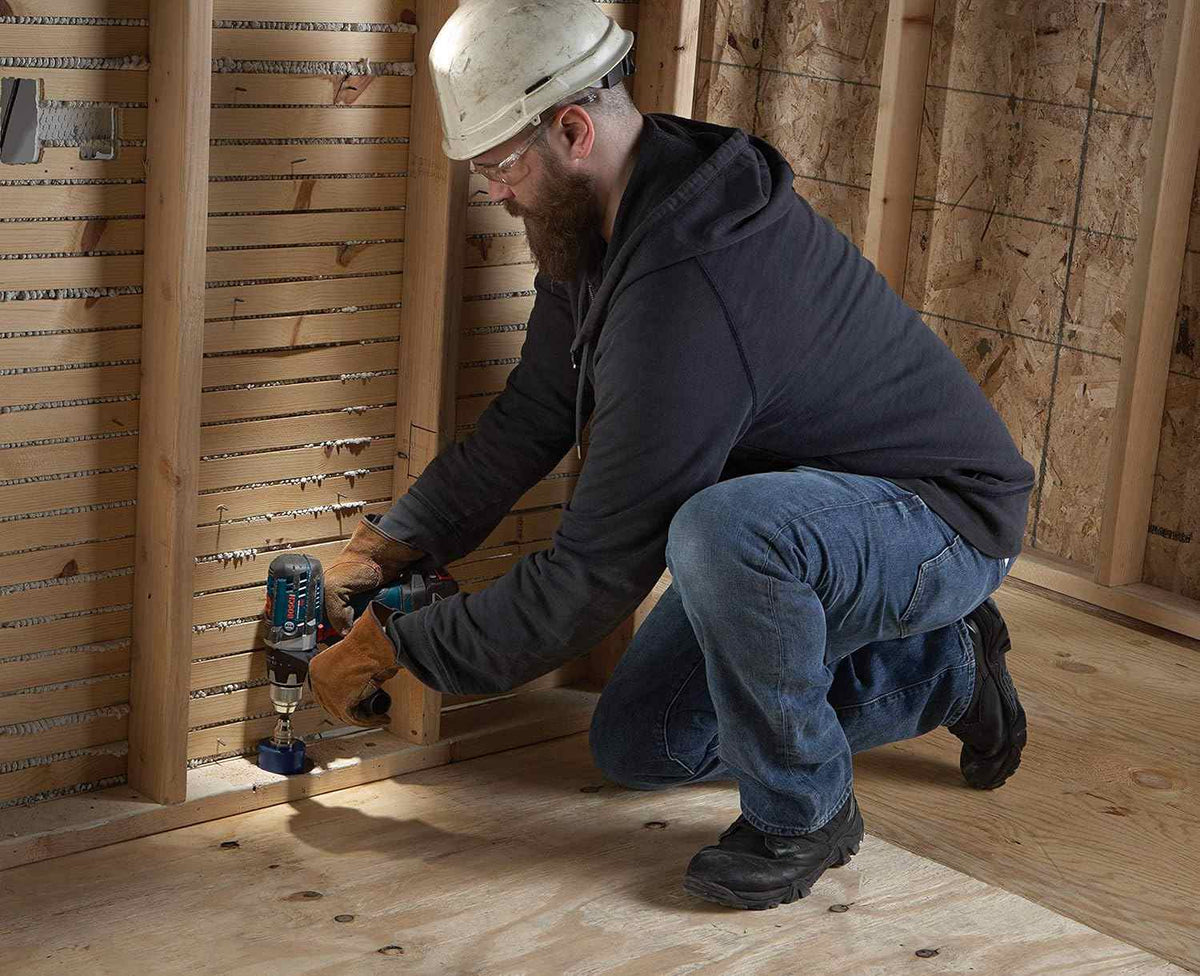 Action shot of a worker using the blue carbide hole saw to drill through the bottom plate of a wooden wall frame.