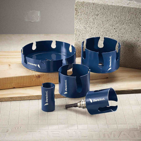 Studio arrangement of various sizes of blue Daredevil carbide hole saws resting on a construction surface next to a concrete block.