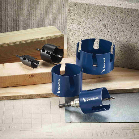 Comparison view showing blue Daredevil carbide hole saws alongside black standard hole saws arranged on a tiled surface with a concrete block.