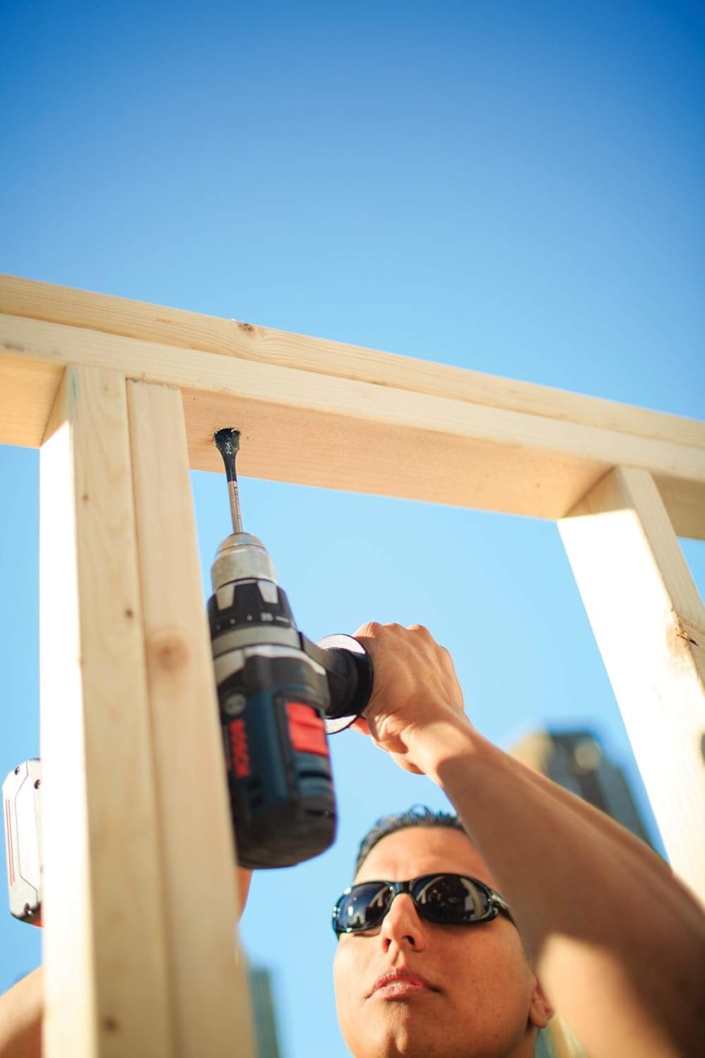 Low angle view of a worker using a drill and spade bit to bore a hole in overhead wooden framing against a blue sky.