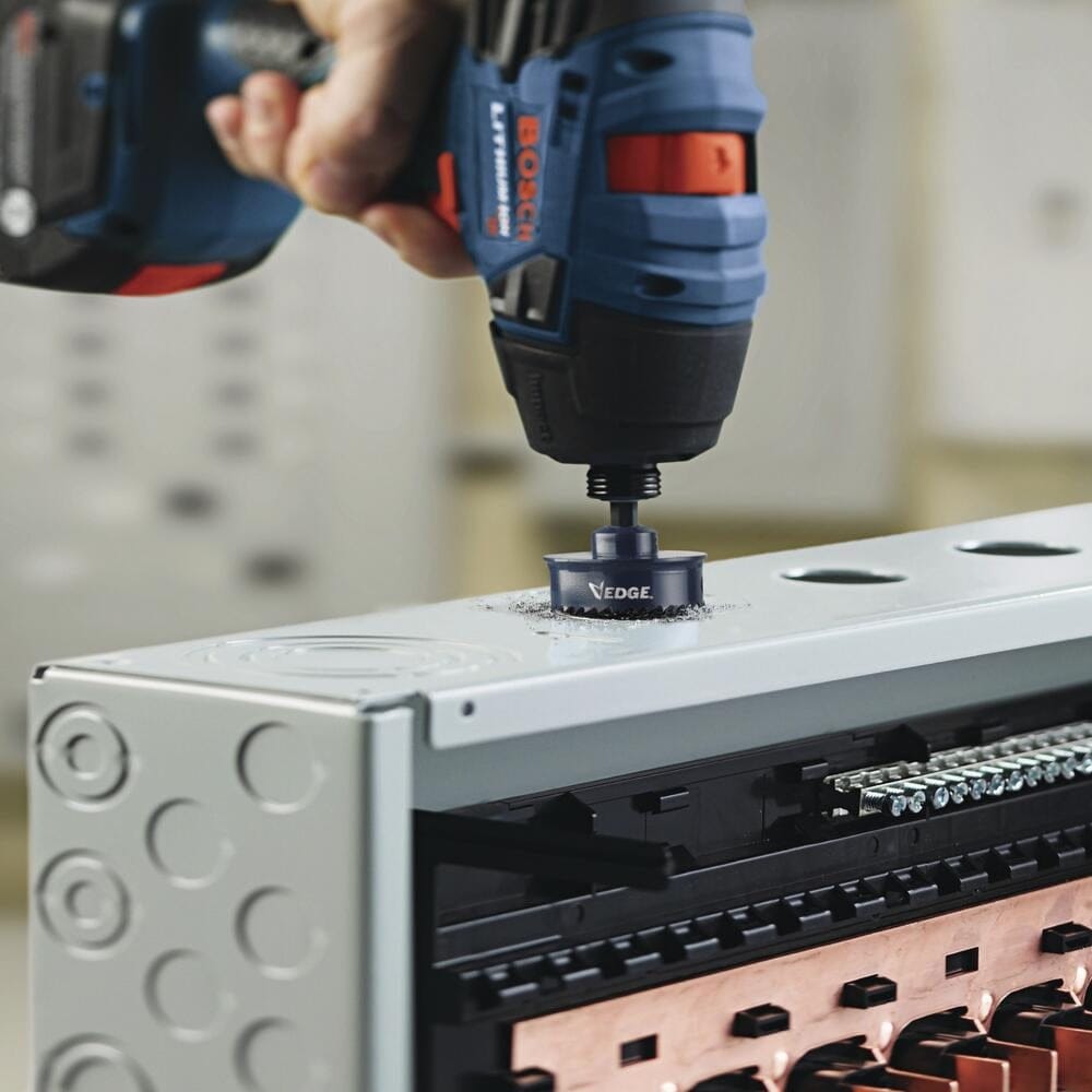 An action photo of a professional using a cordless Bosch impact driver and the 1-1/2 inch HTW112 hole saw to drill a precise knockout hole into a metal electrical enclosure box.