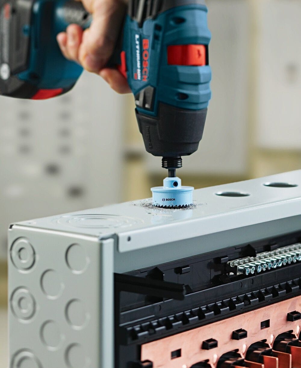 Close-up action shot of a worker using a cordless drill to cut a clean hole through a grey metal electrical box with the hole saw.