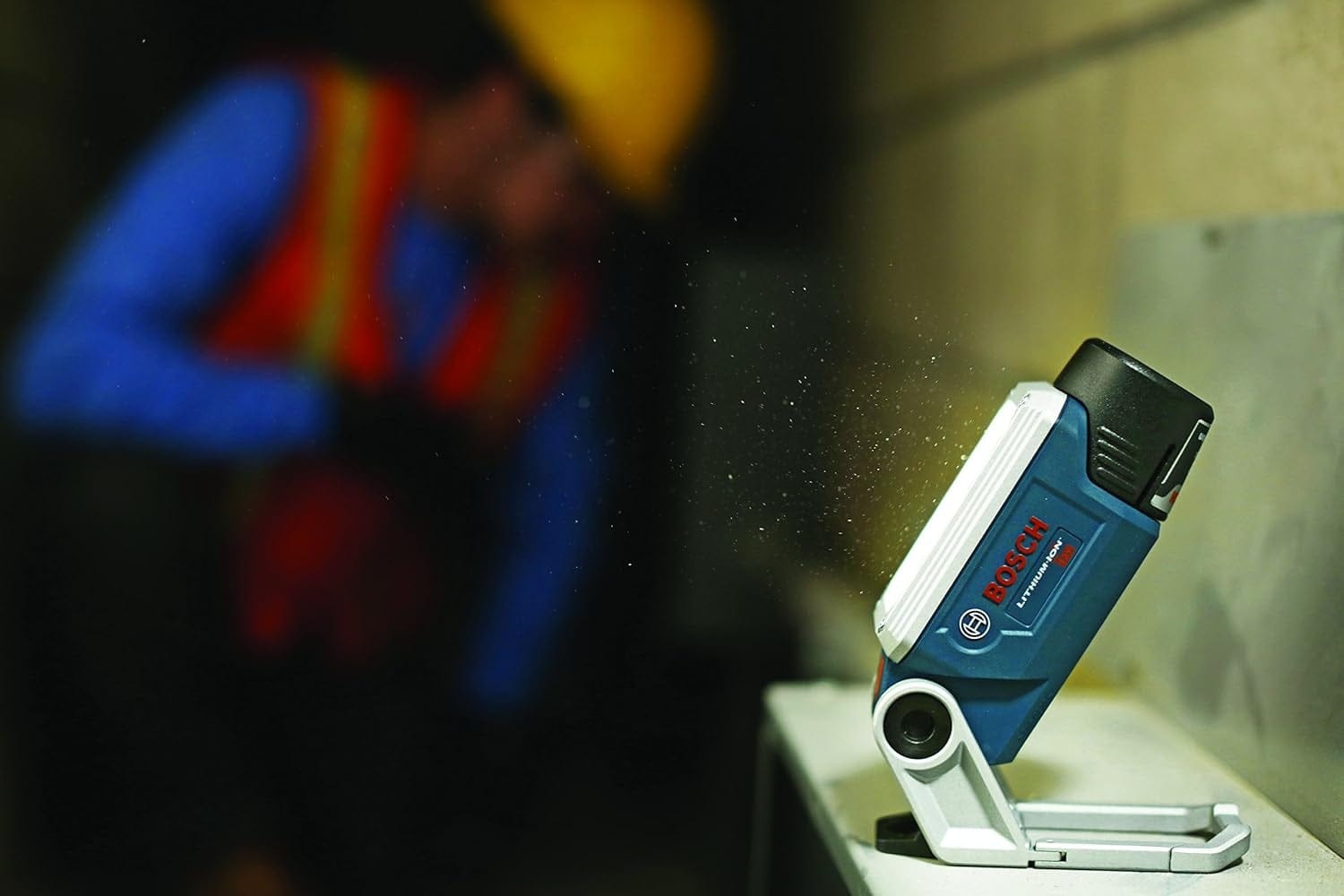 Action shot of the Bosch worklight magnetically secured to a metal surface, illuminating a dark jobsite area with a blurred worker visible in the background.