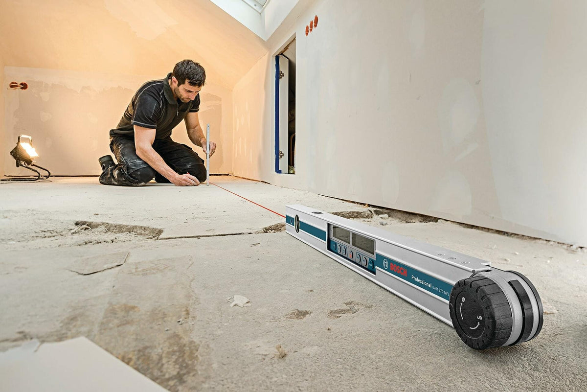 The Bosch GAM 270 MFL rests on a concrete floor, projecting its integrated red laser line for layout, while a worker kneels in the background to mark the line.