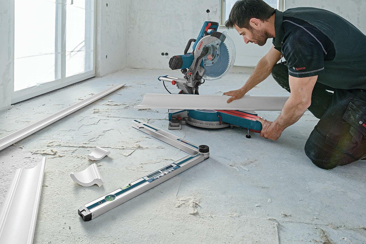A carpenter prepares to cut crown molding on a Bosch miter saw, with the GAM 270 MFL angle finder resting on the floor nearby, illustrating its use in calculating complex miter cuts.
