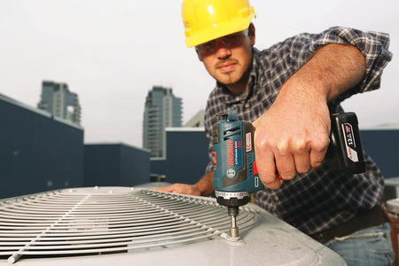 An HVAC professional wearing a hard hat while using a Bosch 12V Max impact driver with a high-capacity battery to service an outdoor ventilation unit.