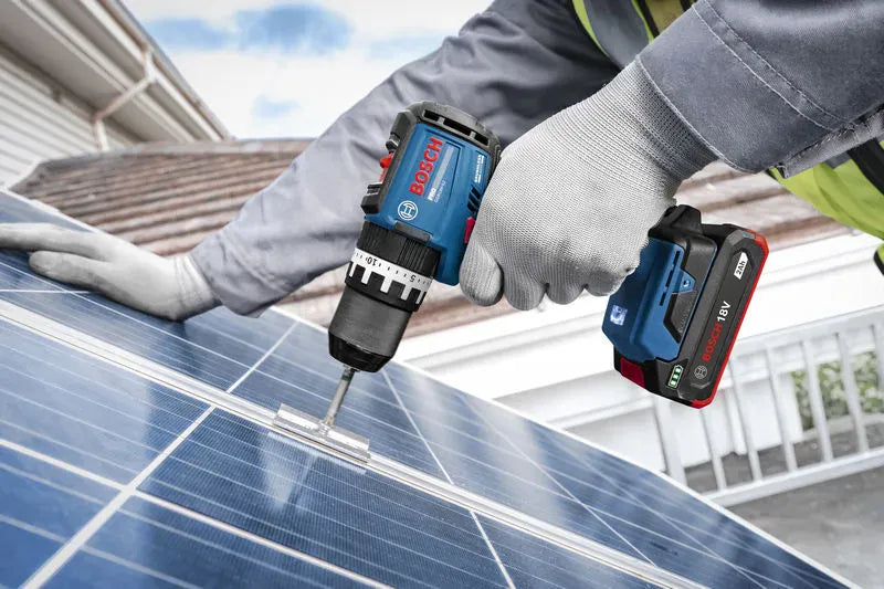 An uncropped real-world field application view showing a rooftop mechanical technician driving fasteners through a solar panel bracket hook run. Equipped with gray safety gloves, the tradesman balances the cordless tool firmly, highlighting the compact, low-profile footprint of the Bosch 18V 2.0Ah battery during demanding outdoor structural setups.