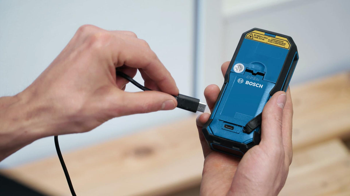 Action shot demonstrating the charging process, with a hand plugging the USB-C cable into the port on the bottom of the laser measure containing the GBA37V10 battery pack