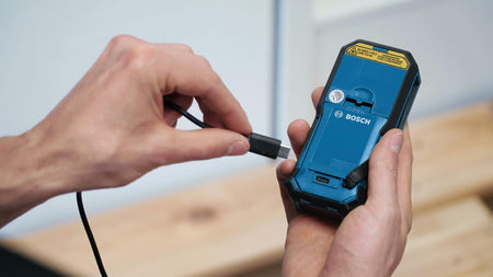 Action shot demonstrating the charging process, with a hand plugging the USB-C cable into the port on the bottom of the laser measure containing the GBA37V10 battery pack