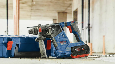 Bosch cordless rotary hammer drill with 18V battery and auxiliary handle placed beside blue carrying case on construction site floor