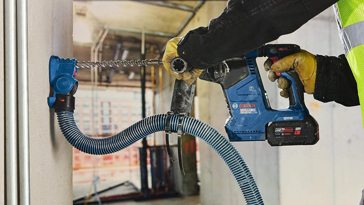Worker wearing safety gear using heavy-duty drill on concrete ceiling; pipes and structural elements visible in background.