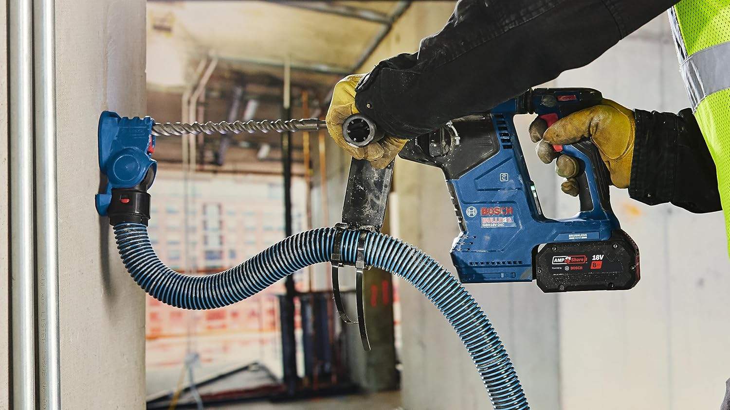 Worker wearing safety gear using heavy-duty drill on concrete ceiling; pipes and structural elements visible in background.