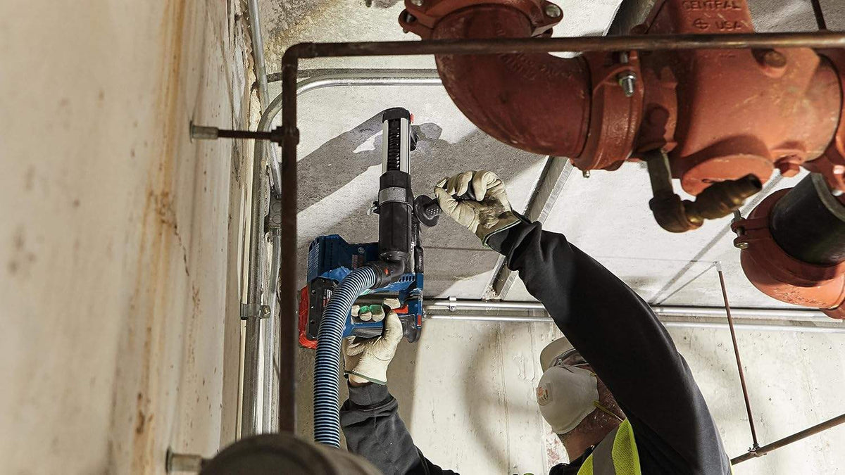 Bosch cordless rotary hammer drill with 18V battery and auxiliary handle placed beside blue carrying case on construction site floor