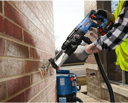 A close-up view of a worker using the Bosch SDS-Plus Bulldog rotary hammer and a HEPA-ready vacuum attachment to perform clean, dustless drilling into an exterior brick wall.