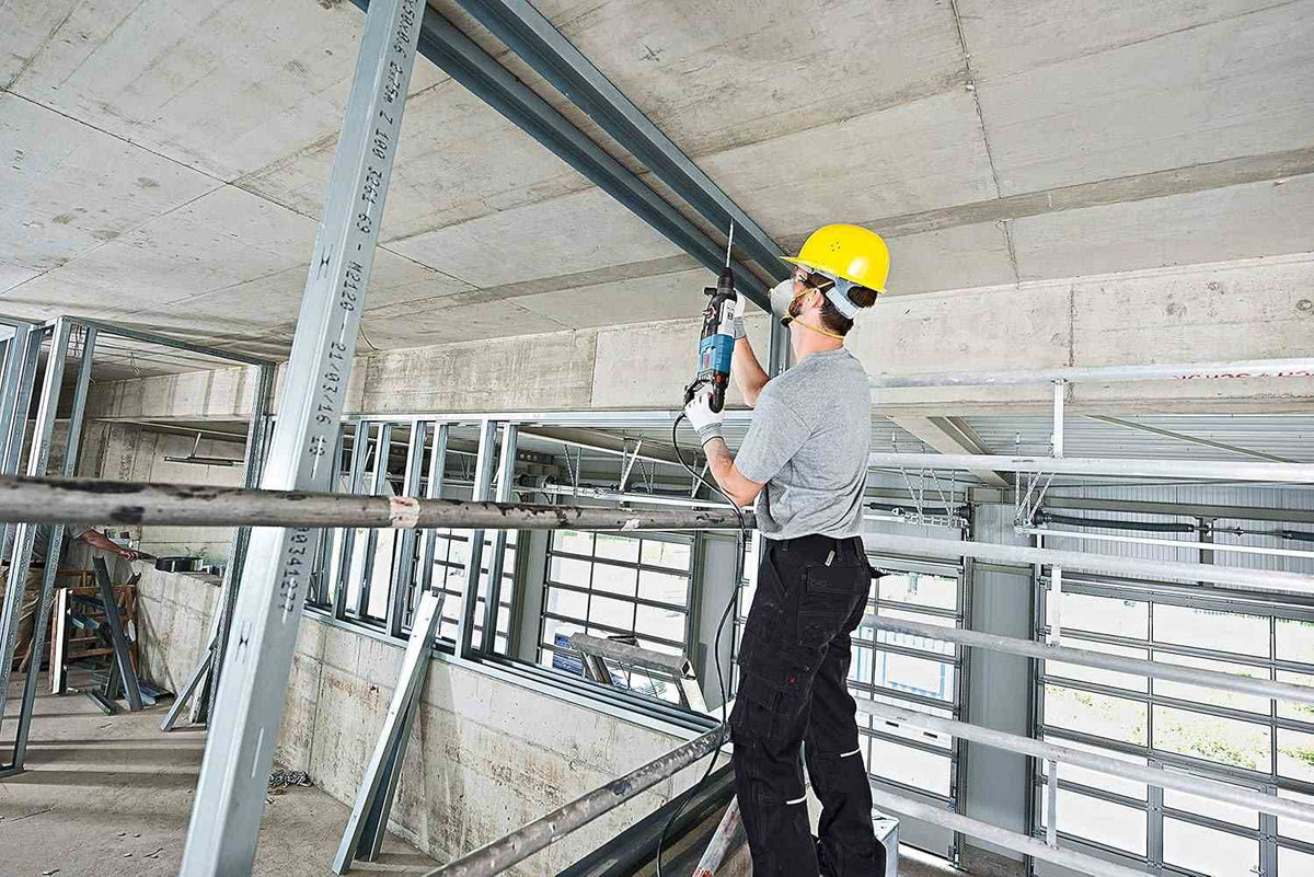 An electrician using the Bosch Bulldog rotary hammer to drill into a steel-reinforced concrete ceiling structure from a scaffold, demonstrating its power and reach.